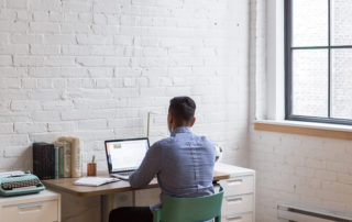 a man wearing a checkered dress shirt sitting at a desk working on a laptop in a bright lit room