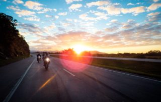 a group of three motorcyclists driving on an empty road with a sunset behind them