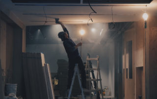 a painter standing on a ladder using a sander on the ceiling leaning backwards with dim lighting