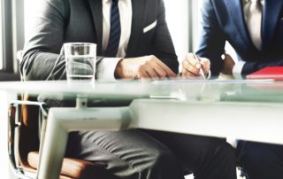 two men wearing suits at a glass table looking over documents