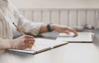 woman sitting at a desk with one hand on a laptop keyboard and the other writing in a notebook