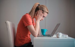 a woman at a desk holding her head in her hands worried looking at a laptop